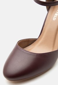 Καφέ δερμάτινο γυναικείο παπούτσι με κλειστή μύτη και λουράκι στον αστράγαλο, με ομαλή υφή και μπεζ εσωτερική επένδυση.