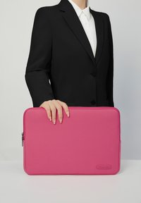 Rosa bærepose til laptop med en glat tekstur, lynlåslukning og logo-detalje. Holdt i en hånd mod en neutral baggrund, parret med en sort blazer.