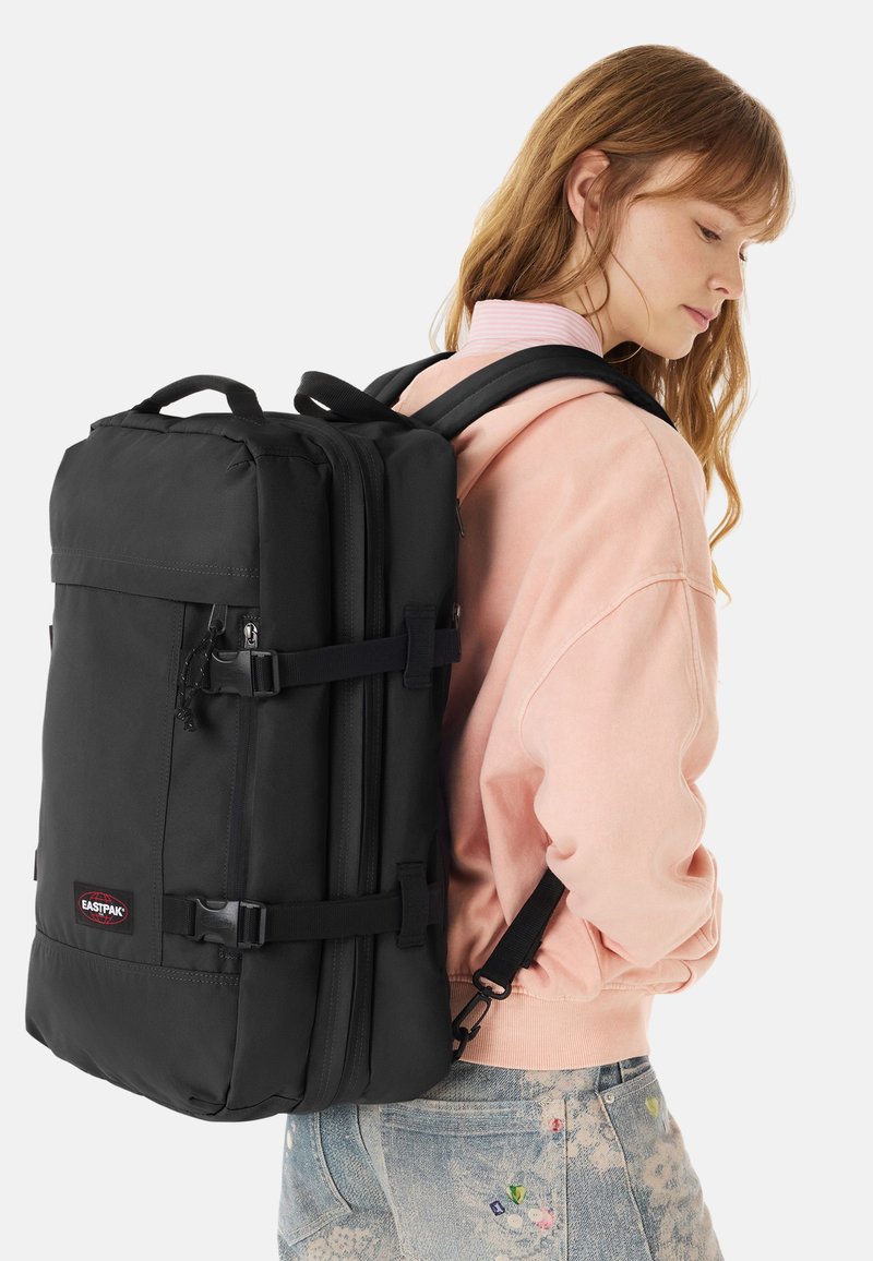 Femme en veste rose portant un grand sac à dos noir Eastpak avec plusieurs compartiments et sangles latérales.