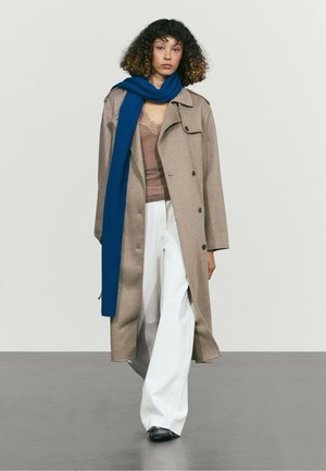 Donna che indossa un cappotto lungo beige, una sciarpa blu, un top marrone, pantaloni bianchi a gamba larga e scarpe nere che cammina su un pavimento grigio con uno sfondo chiaro uniforme.