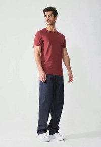 Maglietta maroon a maniche corte, abbinata a jeans di denim blu scuro. L'outfit presenta sneakers bianche e un design a vestibilità rilassata.