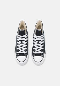 Paire de baskets montantes en toile noire avec embouts en caoutchouc blanc et lacets blancs sur fond clair.