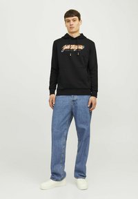 Sudadera negra con un bolsillo canguro en la parte frontal, que presenta un diseño de logo multicolor. Combinada con jeans de mezclilla de pierna recta en un tono azul claro y zapatillas blancas.