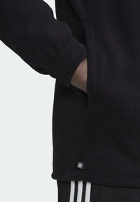 Svart fleece sweatshirt med ribbad mudd, sidofickor och en liten logotyp-patch nära fållen. Slät yta och avslappnad passform.