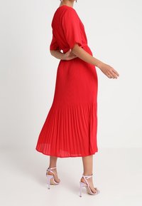 Vestido midi plisado rojo con mangas cortas, que cuenta con una cintura ajustada y una falda fluida. Combinado con unos tacones ligeros de color rosa claro.