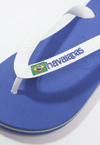 Sinine kummist slaider valgete rihmadega, millel on roheline ja kollane Brasiilia lipu detail ning teksteeritud jalaosa. Nähtav Havaianas logo.