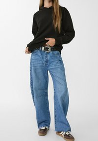 Sweat à capuche noir, jean bleu clair ample avec détails en patchwork, ourlet effiloché, et baskets marron. Ceinture argentée avec boucle ornée.