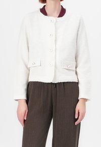 Biała kurtka o fakturze z okrągłym kołnierzem, zapięciem na guziki z przodu oraz dwiema kieszeniami naszytymi. Posiada kontrastujący bordowy wewnętrzny kołnierz.