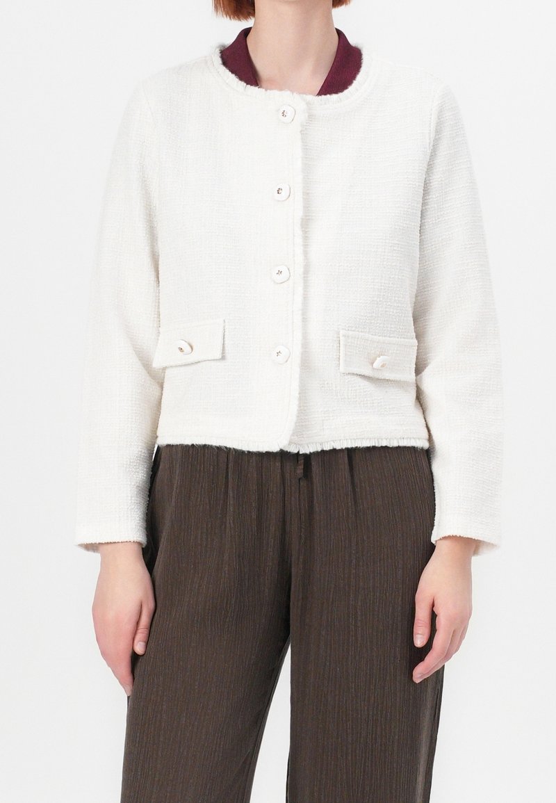 Biała kurtka o fakturze z okrągłym kołnierzem, zapięciem na guziki z przodu oraz dwiema kieszeniami naszytymi. Posiada kontrastujący bordowy wewnętrzny kołnierz.