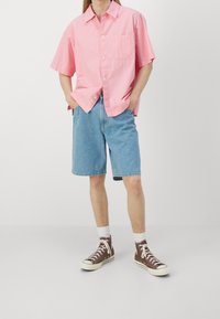 Ljusrosa kortärmad skjorta med knappar och bröstficka, i kombination med blå denimshorts och bruna högklackade sneakers med vita sulor.