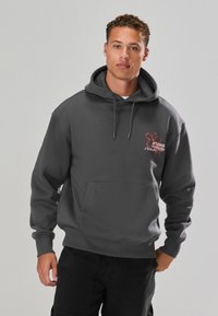 Felpa con cappuccio grigia in tessuto morbido, dotata di tasca frontale e cappuccio con cordino regolabile. Logo grafico rosso ben visibile sul petto.