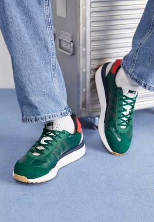 Personne portant des baskets vertes, blanches et rouges avec des lacets vert foncé et un jean bleu clair, debout près d'une mallette en métal argenté sur un sol bleu.