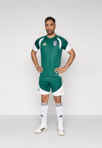 Farda de futebol verde feita de tecido de malha, com detalhes brancos, dourados e o emblema da Itália. Calções com bainha branca em contraste, combinados com meias brancas.