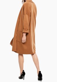 Hnědý pletený oversized cardigan se šálovým límcem, tříčtvrtečními rukávy a předními kapsami; nošený přes přiléhavé černé šaty a podpatky.