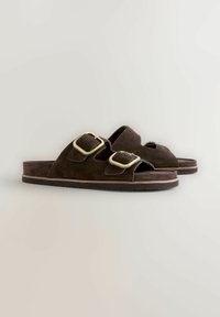 Sandalias abiertas de ante marrón con dos grandes hebillas doradas y suelas de goma planas sobre un fondo claro y liso.