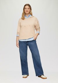 Helle beige Kapuzenpullover über einem hellblauen Hemd, kombiniert mit lockeren blauen Jeans und beigen Sneakers mit dunklen Akzenten.
