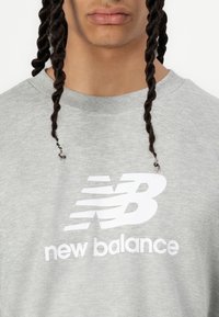 Világosszürke pulóver, amelyen egy kiemelkedő fehér "New Balance" logó található, sima felülettel és bordázott kerek nyakkivágással.