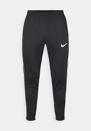 Black athletic pants with elastic waistband, tapered legs, gold Nike logo on right side, and white stripes along the sides. Soft, smooth fabric.