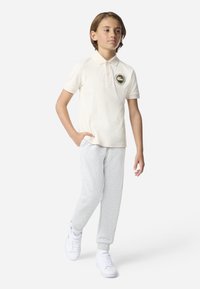 Kremfarget poloshirt med logo, kombinert med lysegrå joggebukser og hvite joggesko. Poloshirten har korte ermer og klassisk krage.