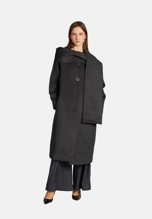Abrigo de lana gris oscuro con un diseño estructurado, gran cuello y cinco botones negros. Acompañado de pantalones de pierna ancha en negro y tacones.