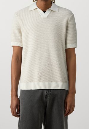 Person trägt ein weißes, strukturiertes, kurzärmeliges Strick-Poloshirt mit gerippten Bündchen und Saum, kombiniert mit dunkelgrauen Hosen.
