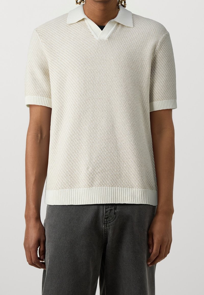 Personne portant un polo en maille blanche texturée à manches courtes avec poignets et ourlet côtelés, associé à un pantalon gris foncé.