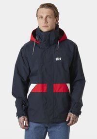 Marineblauwe waterdichte jas met een ontwerp van rode en witte strepen, hoge kraag, verstelbare capuchon en zilverkleurige hardware. Bevat een logo op de borst.