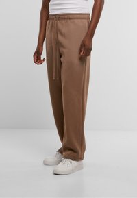 Braune, weiche Stoff-Sweatpants mit einem elastischen Bund und Kordelzug, die eine lockere Passform und Seitentaschen bieten, kombiniert mit weißen Turnschuhen.