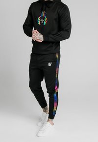 Czarna bluza z kapturem z kolorową grafiką, czarne dresy z tęczowym paskiem, i białe trampki. Materiał wydaje się gładki i elastyczny.