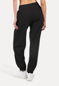 Černé tepláky s elastickým pasem a manžetami, vyrobené z měkkého materiálu, s volným střihem. Nosič má na sobě bílé sportovní boty.