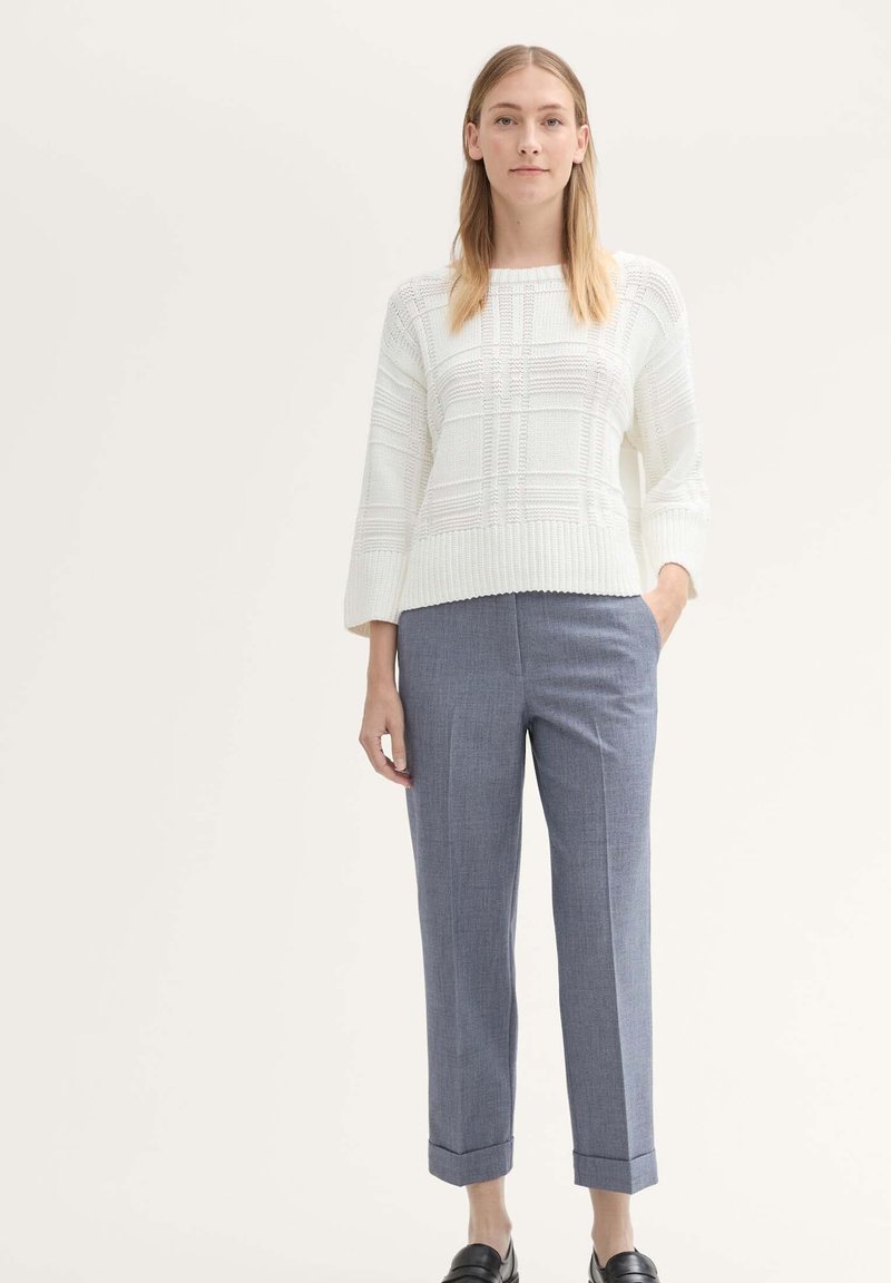 Witte gebreide trui met een gestructureerd ruitenpatroon, gecombineerd met lichtblauwe op maat gemaakte broek en zwarte loafers. Model staat tegen een neutrale achtergrond.