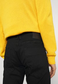 Jeans negri dintr-un material texturat, cu buzunare tip patch și detalii de cusătură vizibile, asortate cu un pulover galben din tricot.