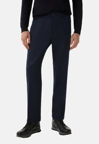 Granatowe spodnie o klasycznym kroju, z przodu z zakładkami, elastycznym pasem i zapięciem na guzik. Zestawione z czarnymi sportowymi butami.