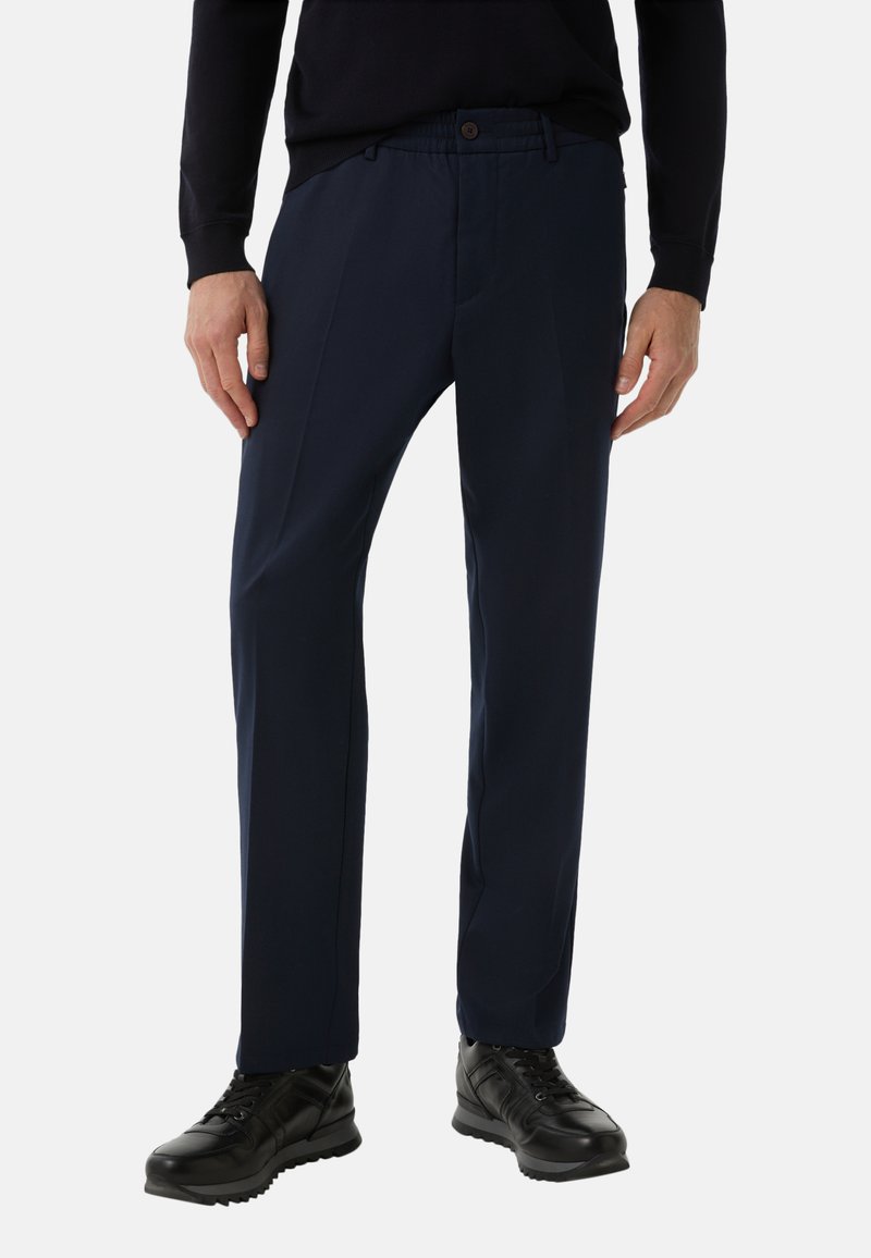 Granatowe spodnie o klasycznym kroju, z przodu z zakładkami, elastycznym pasem i zapięciem na guzik. Zestawione z czarnymi sportowymi butami.