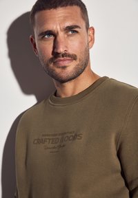 Mand med kort hår og skæg iført en olivengrøn sweatshirt med teksten "Crafted Goods", der kigger let til siden mod en lys baggrund.