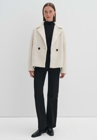 Weißer, strukturierter, doppelt geknöpfter Außenjacke mit großen schwarzen Knöpfen, kombiniert mit einem schwarzen gerippten Rollkragenpullover und engen schwarzen Hosen.