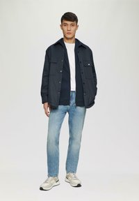 Veste légère marine foncée sur un T-shirt crème, associée à un jean bleu clair et des sneakers beige. La veste dispose de poches avant et d'un col.