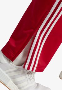 Rote Sporthosen mit weißen Streifen und einem Reißverschluss am Saum, kombiniert mit weißen Sportschuhen mit strukturierten Details und Schnürsenkeln.