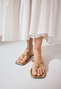 Goldene flache Sandalen mit gebundenem Riemendesign, quadratischer Zehenform und glattem Lederfinish, kombiniert mit einem fließenden weißen Rock.