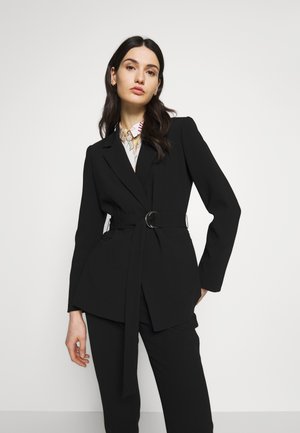 Blazer negro entallado con cintura ceñida, solapas afiladas y mangas largas. La tela parece suave, complementada por una camisa con estampado debajo.