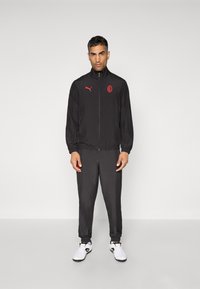 Črna zip-up jakna z rdečimi detajli, usklajene črne hlače z elastičnimi manšetami in logotipom na prsih. Obuta z belimi športnimi copati.