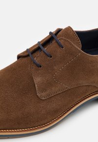 Chaussure de cérémonie en daim marron avec une texture lisse, comportant des lacets bleus, un bout arrondi et une semelle claire contrastante avec des détails de couture.