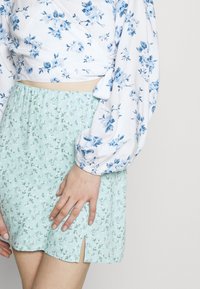 Bílá blůza s modrým květinovým potiskem a nadýchanými rukávy, kombinovaná s světle mátově zelenou sukní s malým rozparkem a jemným květinovým vzorem.