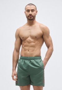 Πράσινα μαγιό σορτς με ελαστική μέση, πλαϊνές τσέπες, πλαϊνές λεπτομέρειες και απαλή υφή, φορεμένα από μοντέλο με καλογυμνασμένο σώμα.