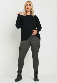Suéter negro de punto acanalado combinado con pantalones cargo verde oliva que tienen bolsillos y botas de combate negras, de pie frente a un fondo neutro.