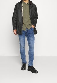 Černá kožená bunda s hnědým límcem, zelená košile na knoflíky, modré slim-fit džíny a černé šněrovací boty.