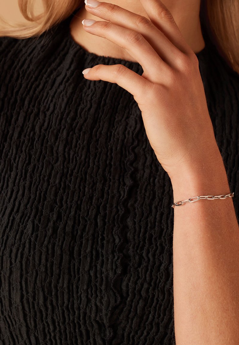 Zilveren schakelsieraad op een arm met een lichte huidtint, rustend tegen een gestructureerde zwarte stof met een golvend patroon en een subtiele glans.