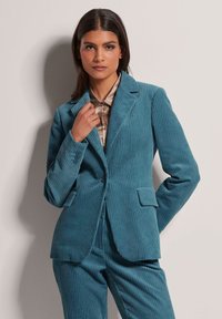 Teal corduroy pak met verticale strepen. Het colbert heeft een revers, voorzakken en een knoopsluiting. Het model draagt een lichtgekleurde blouse eronder.