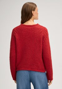 Maglione rosso lavorato a maglia con un design testurizzato a costine e scollo rotondo. Presenta maniche lunghe e una vestibilità rilassata, abbinato a jeans blu.