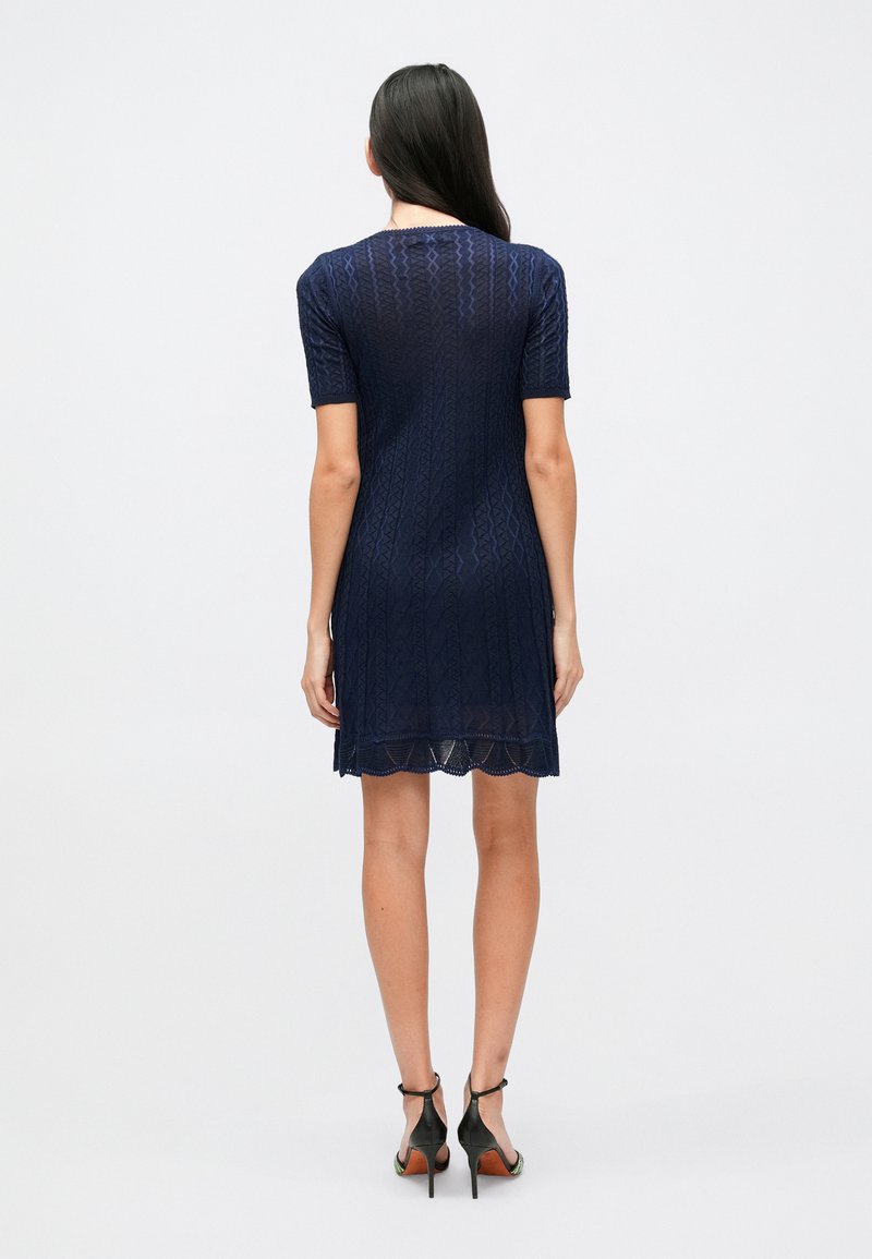 Navy blue knit dress with short sleeves, featuring a textured pattern and scalloped hemline. Paired with black high-heeled shoes.